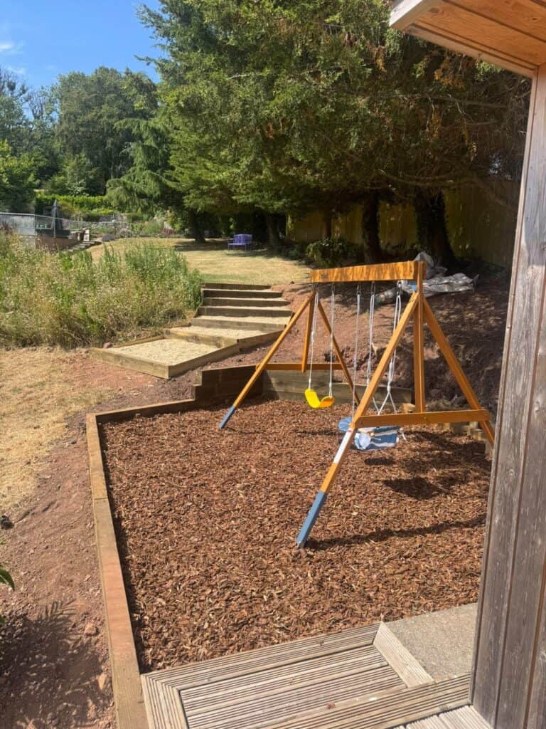 Child's swing set in a garden with trees and steps in the background.
